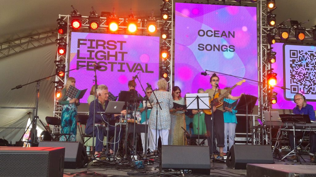 Nic Pendlebury, Lottie P and Colin Riley performing onstage in front of a community choir.
