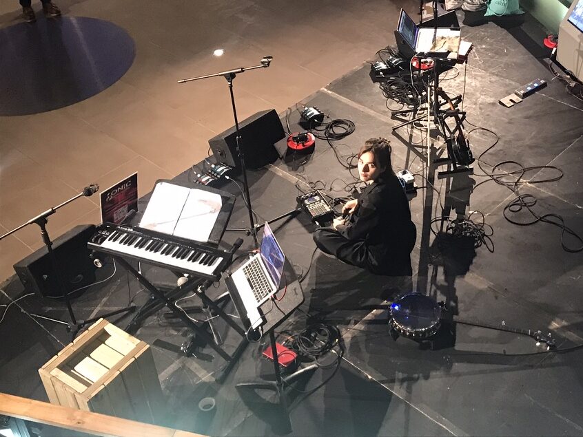 The musician Lottie P (featured on Ebb and Flow) sitting on stage, surrounded by equipment, looking over her shoulder at the camera.